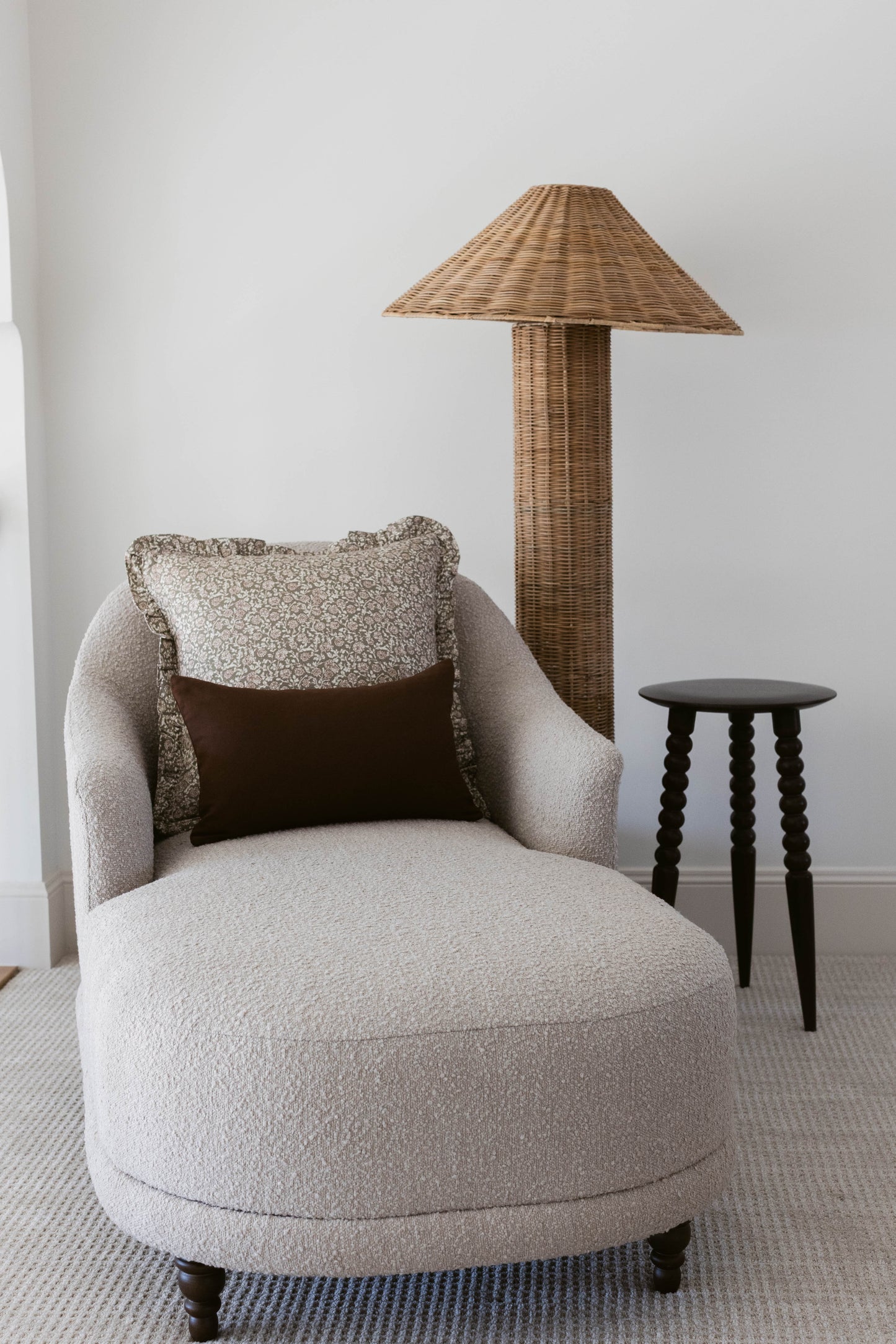Cream upholstered curved armchair with black pillow, woven rattan floor lamp, and black side table in neutral interior settin