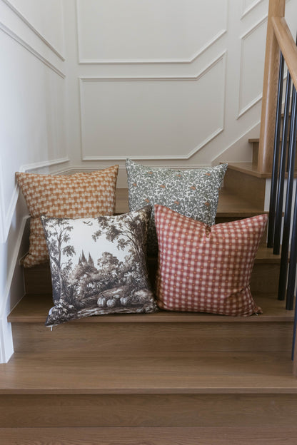 Four decorative pillows with checkered and botanical patterns in tan, gray, and red displayed on wooden stairs.