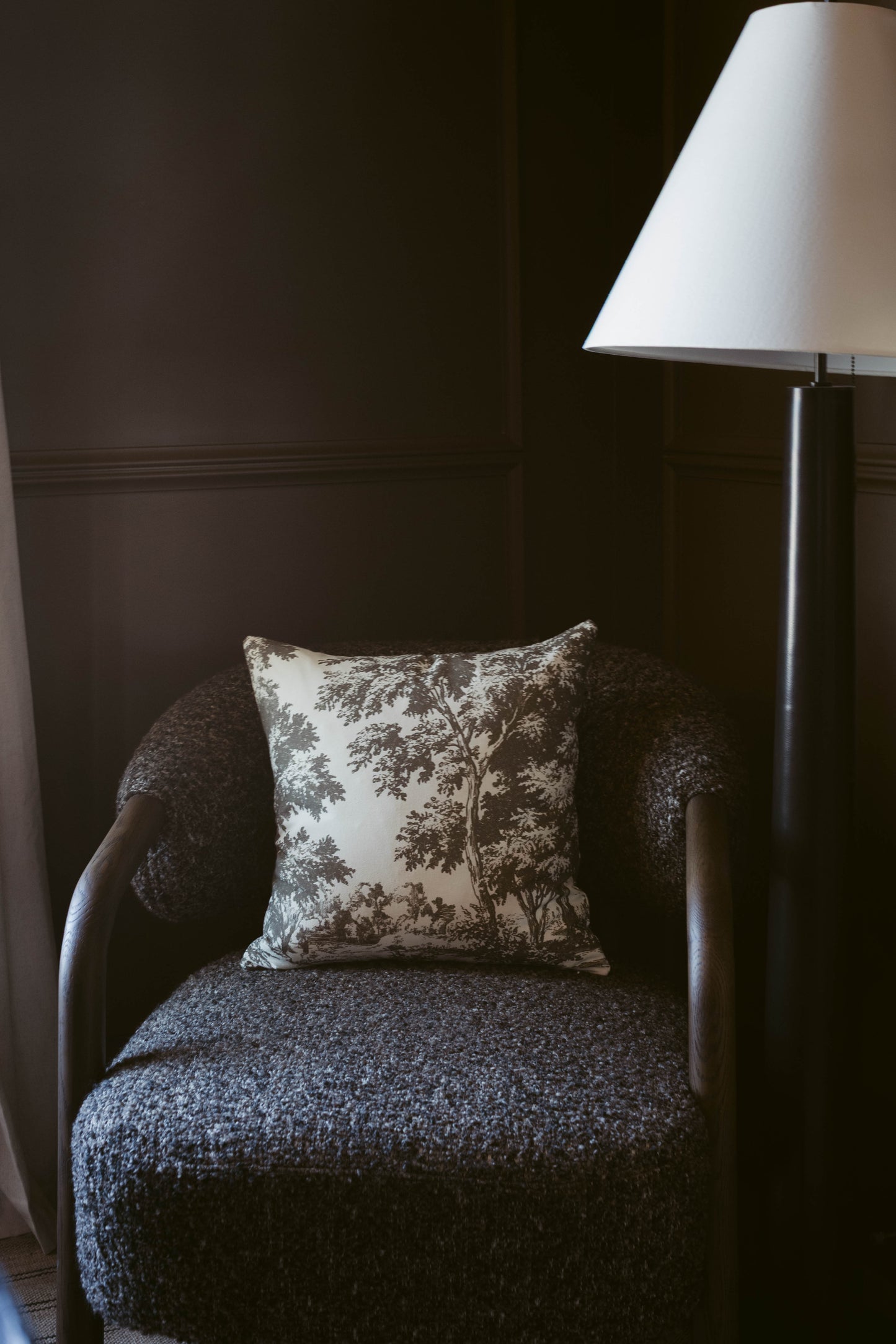 Gray upholstered armchair with tree-print pillow and floor lamp with white shade against dark wall.