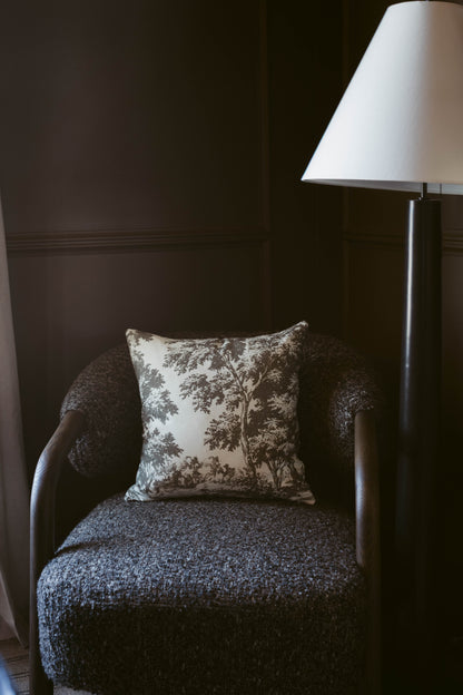 Gray upholstered armchair with tree-print pillow and floor lamp with white shade against dark wall.