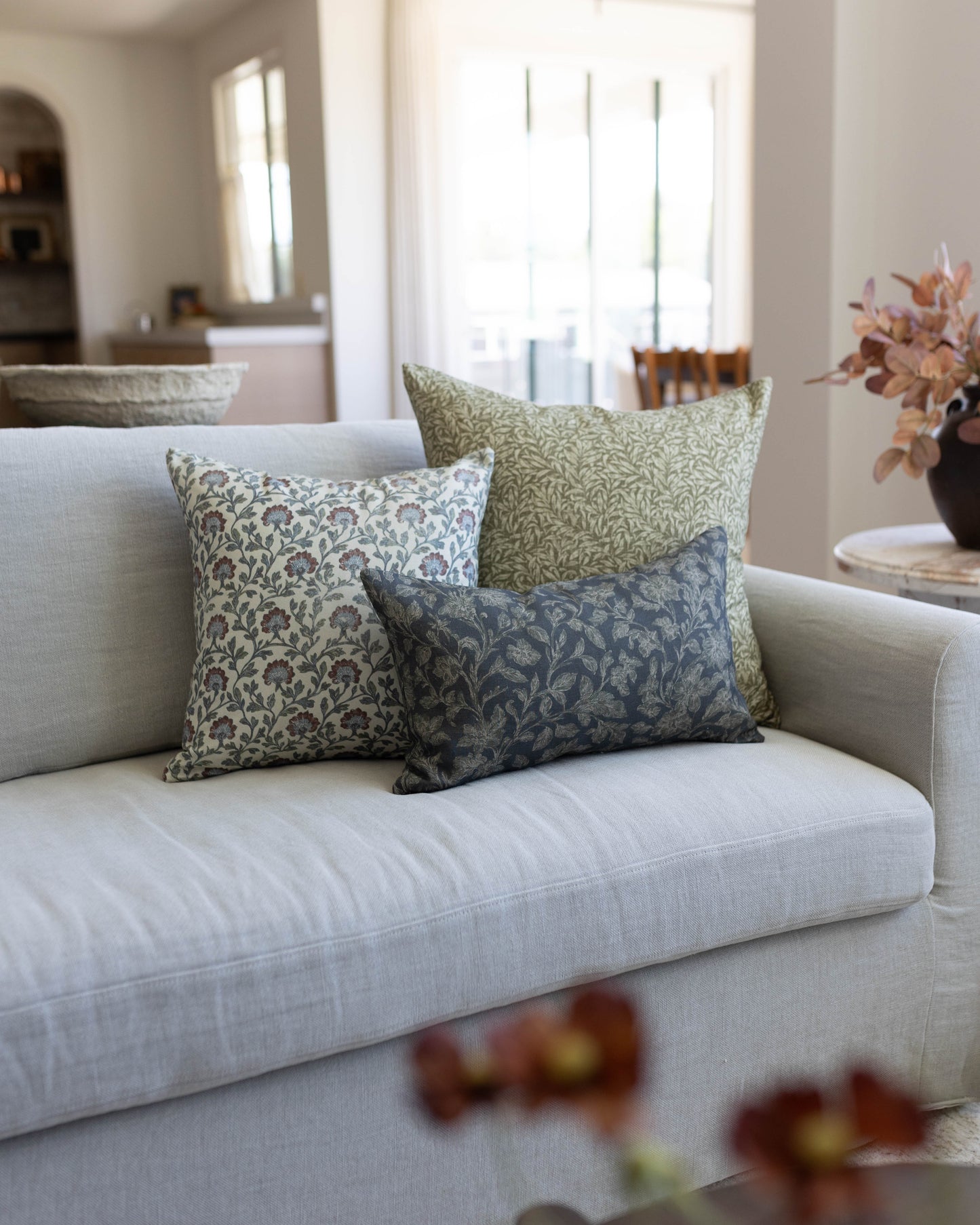 Three decorative pillows with multicolor leaf patterns in cream, green, and navy displayed on a light gray sofa.