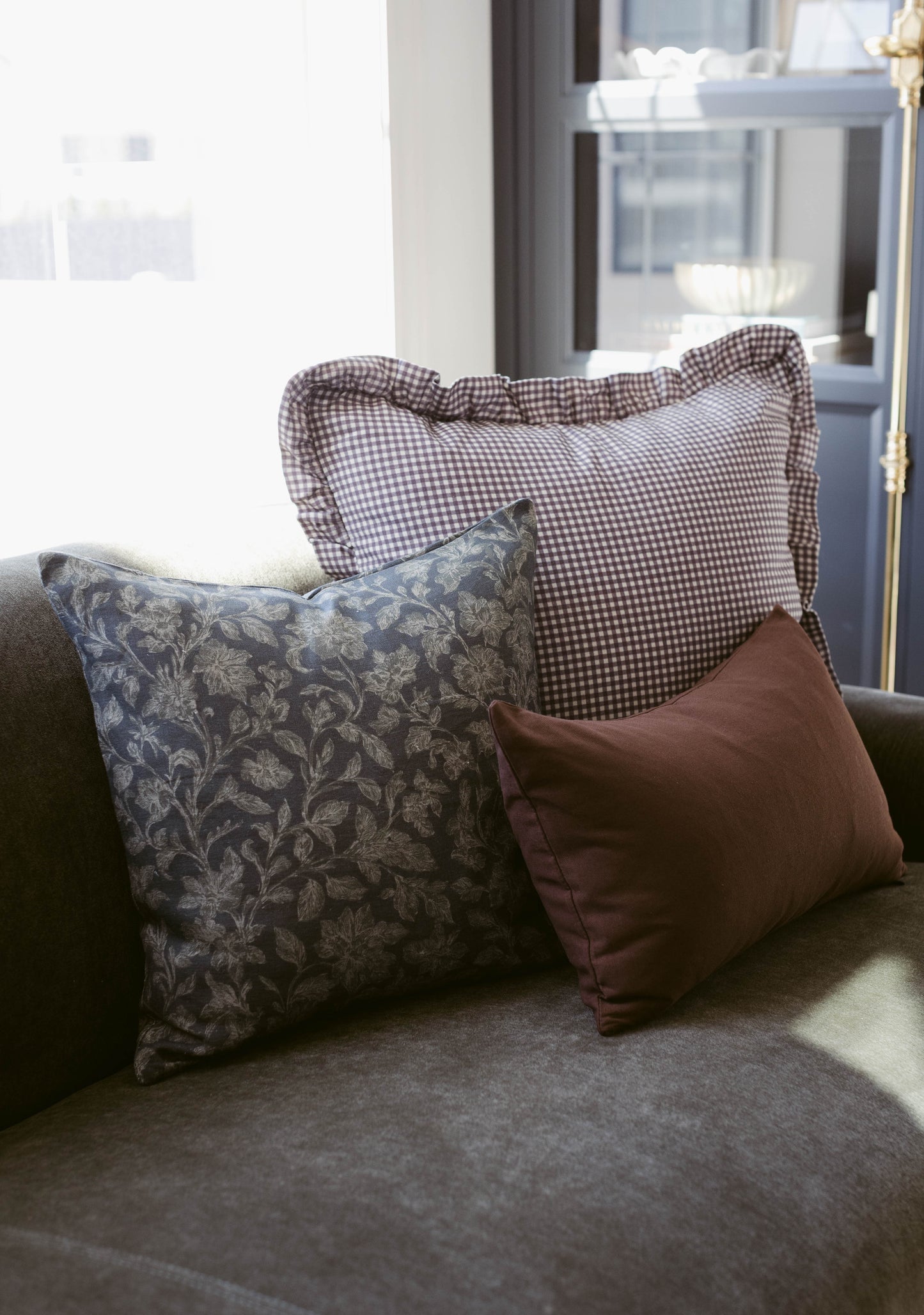 Three decorative pillows arranged on a gray upholstered chair: one with dark blue floral pattern, one with brown gingham chec
