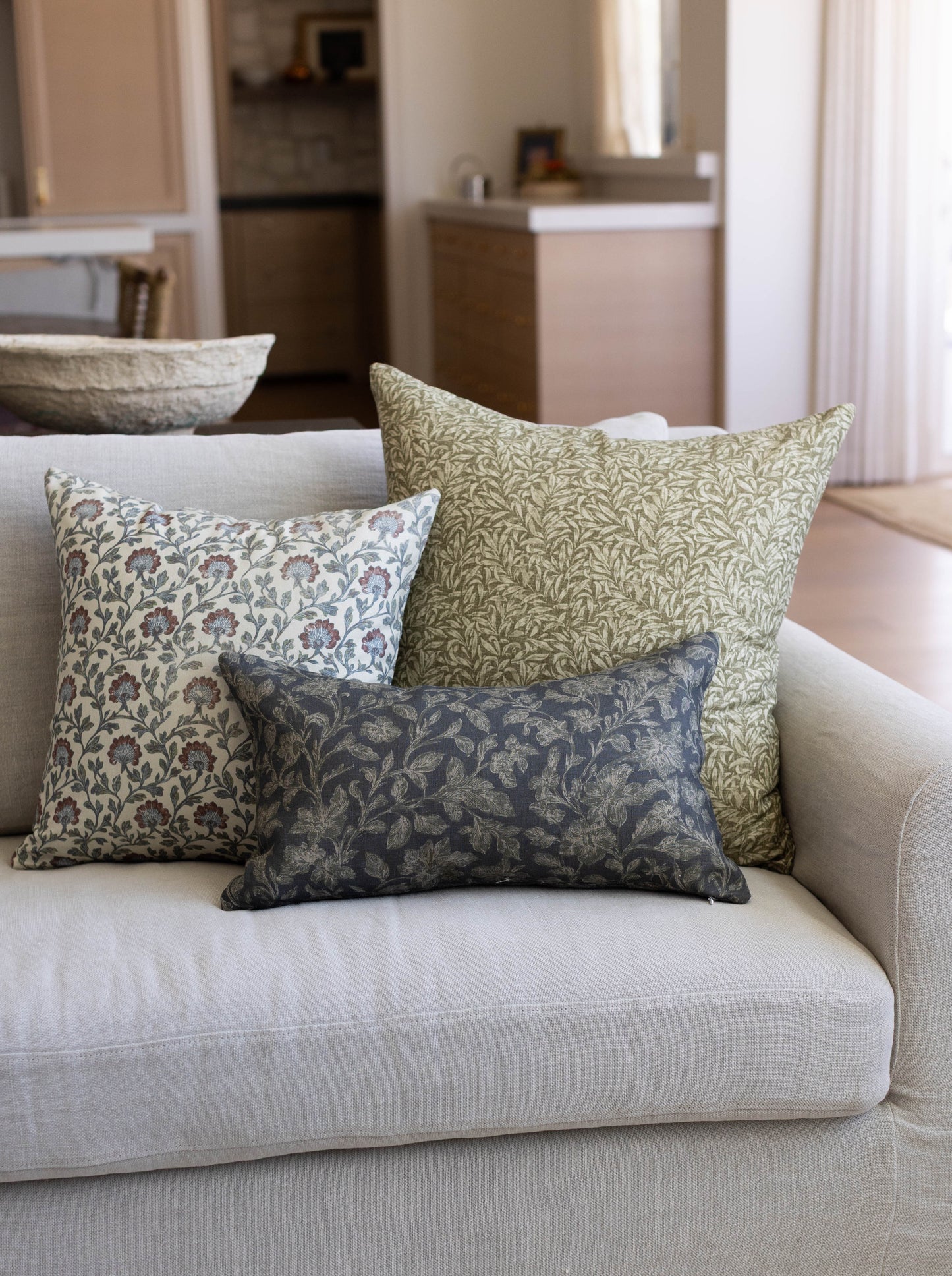 Three decorative pillows with floral and leaf patterns in cream, blue, and sage green on a beige sofa.
