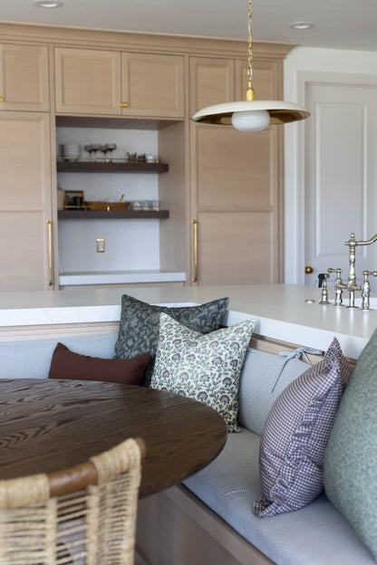 Modern kitchen with beige cabinetry, white countertop, brass-accented pendant light, and banquette seating with patterned pil