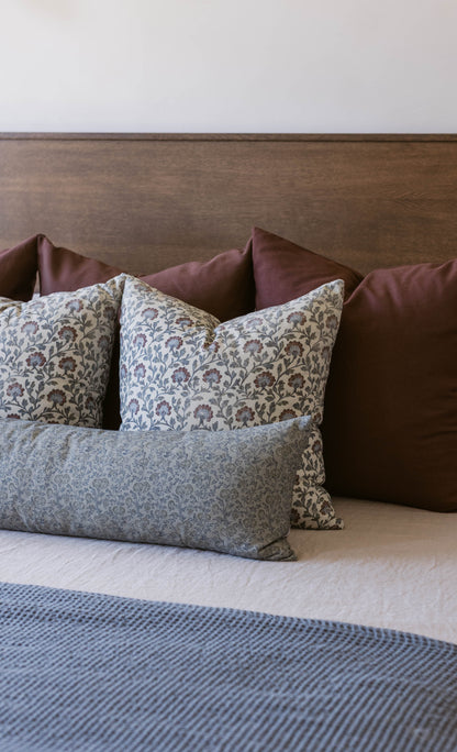 Decorative pillows with multicolor floral and botanical patterns arranged on a bed with burgundy headboard.