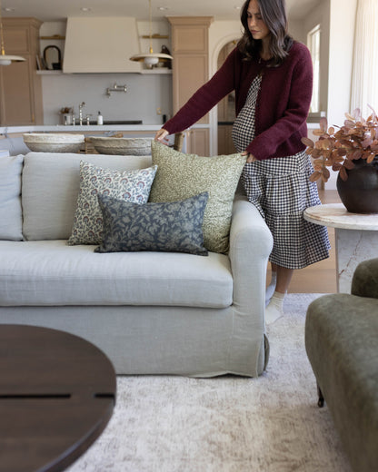 Woman arranging patterned throw pillows on a cream linen sofa in a living room.