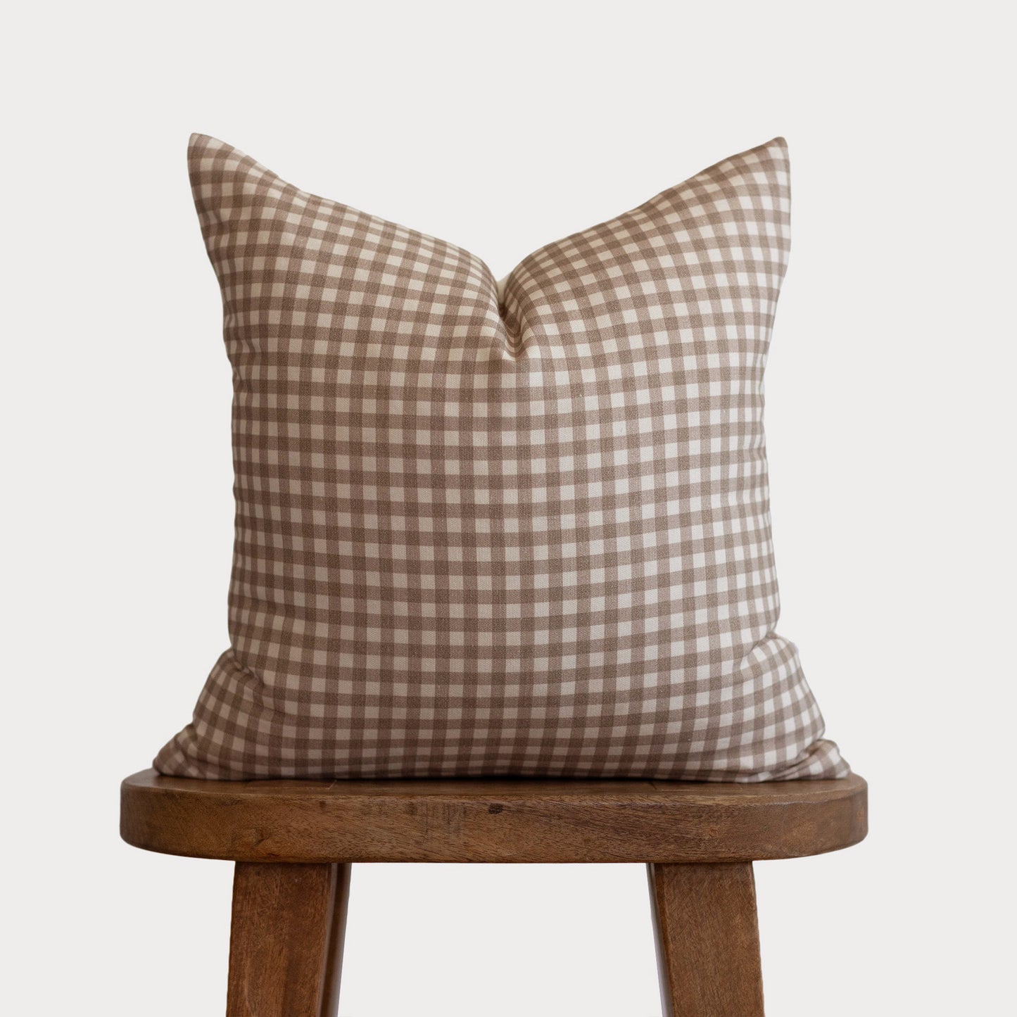 Tan and white gingham plaid square pillow with knife edges displayed on a wooden stool.