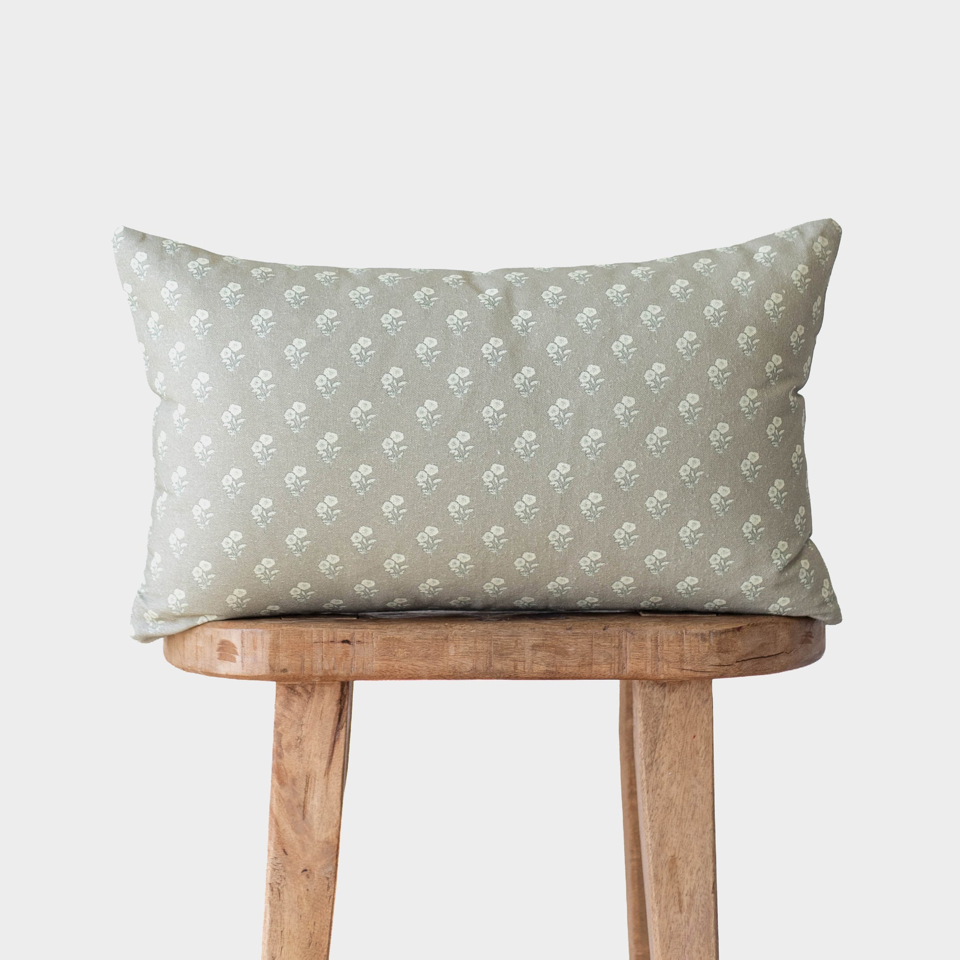 Sage green pillow with white floral pattern resting on wooden stool.
