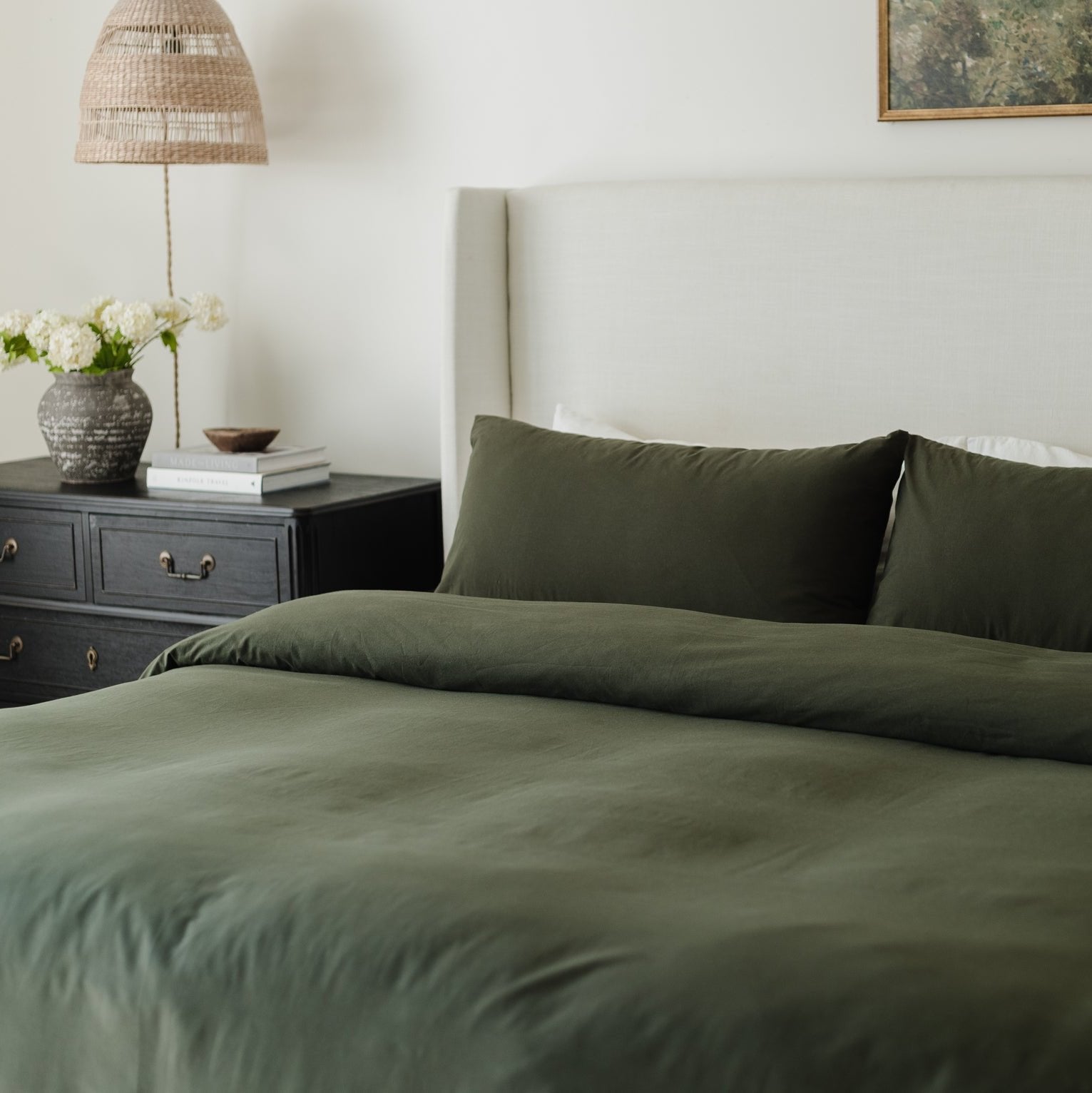 Olive green duvet cover and pillowcases on a made bed next to a nightstand with lamp and flowers.