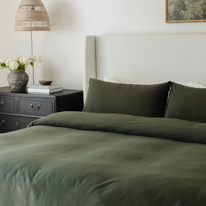 Olive green duvet cover and pillowcases on a made bed next to a nightstand with lamp and flowers.
