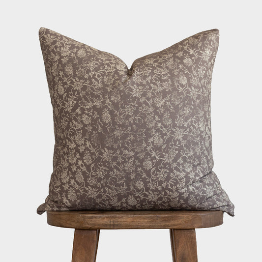 Taupe floral patterned pillow with cream botanical design, displayed on wooden chair legs.