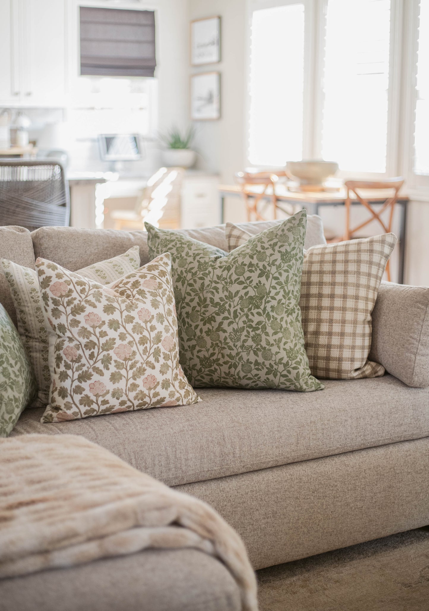 Five decorative throw pillows with floral and checkered patterns arranged on a beige sofa in a bright living room.