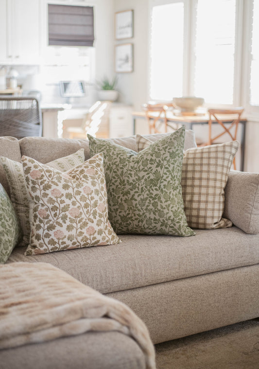 Five decorative throw pillows with floral and checkered patterns arranged on a beige sofa in a bright living room.