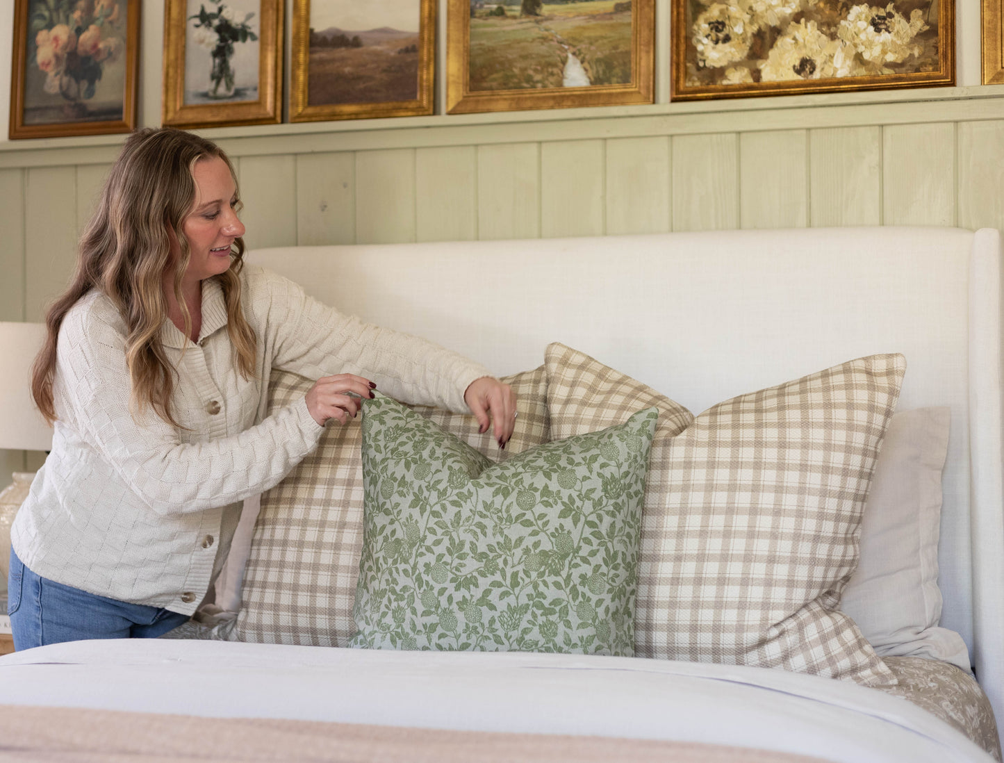 Woman displaying green and beige patterned cotton throw pillows on a bed with white headboard.