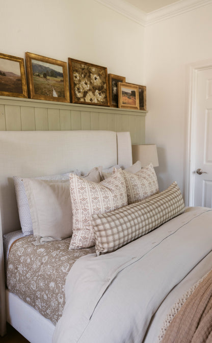 Bed styled with white upholstered headboard, neutral bedding, and decorative pillows in cream, tan, and checkered patterns.