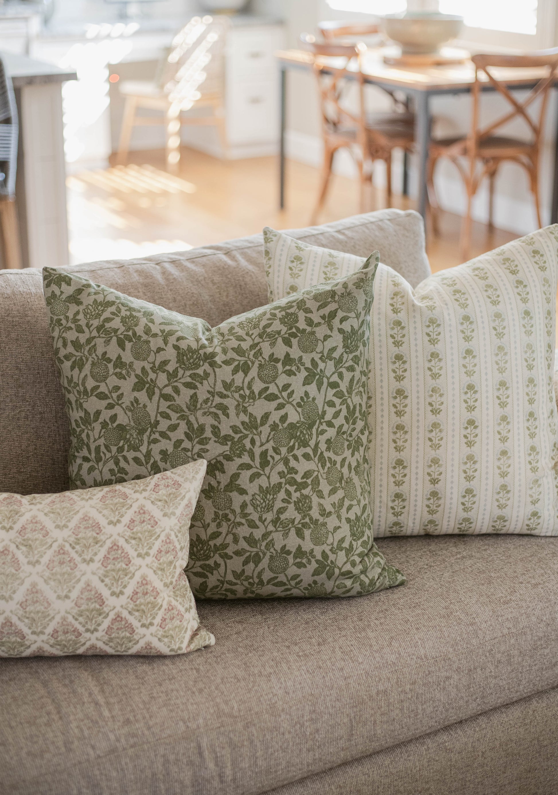Five decorative pillows with green floral and striped patterns arranged on a gray sofa in a bright living room.