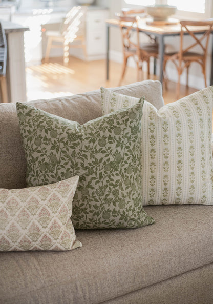 Five decorative pillows with green floral and striped patterns arranged on a gray sofa in a bright living room.