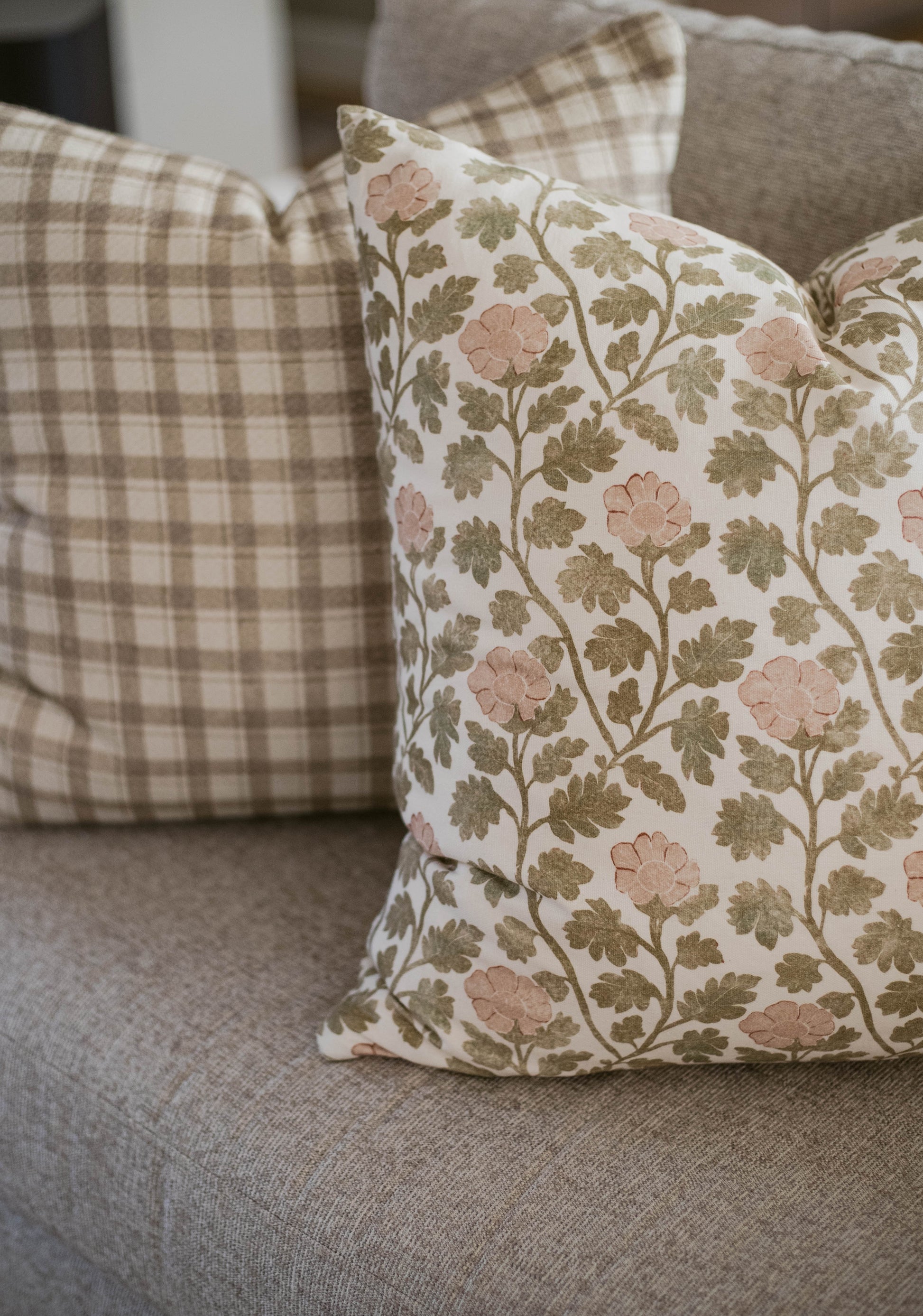 Five decorative pillows with floral and plaid patterns in cream, blush, and sage tones arranged on a gray upholstered sofa.