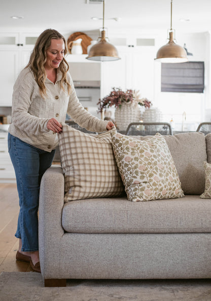 Woman arranging five decorative throw pillows with plaid and floral patterns on a gray upholstered sofa.