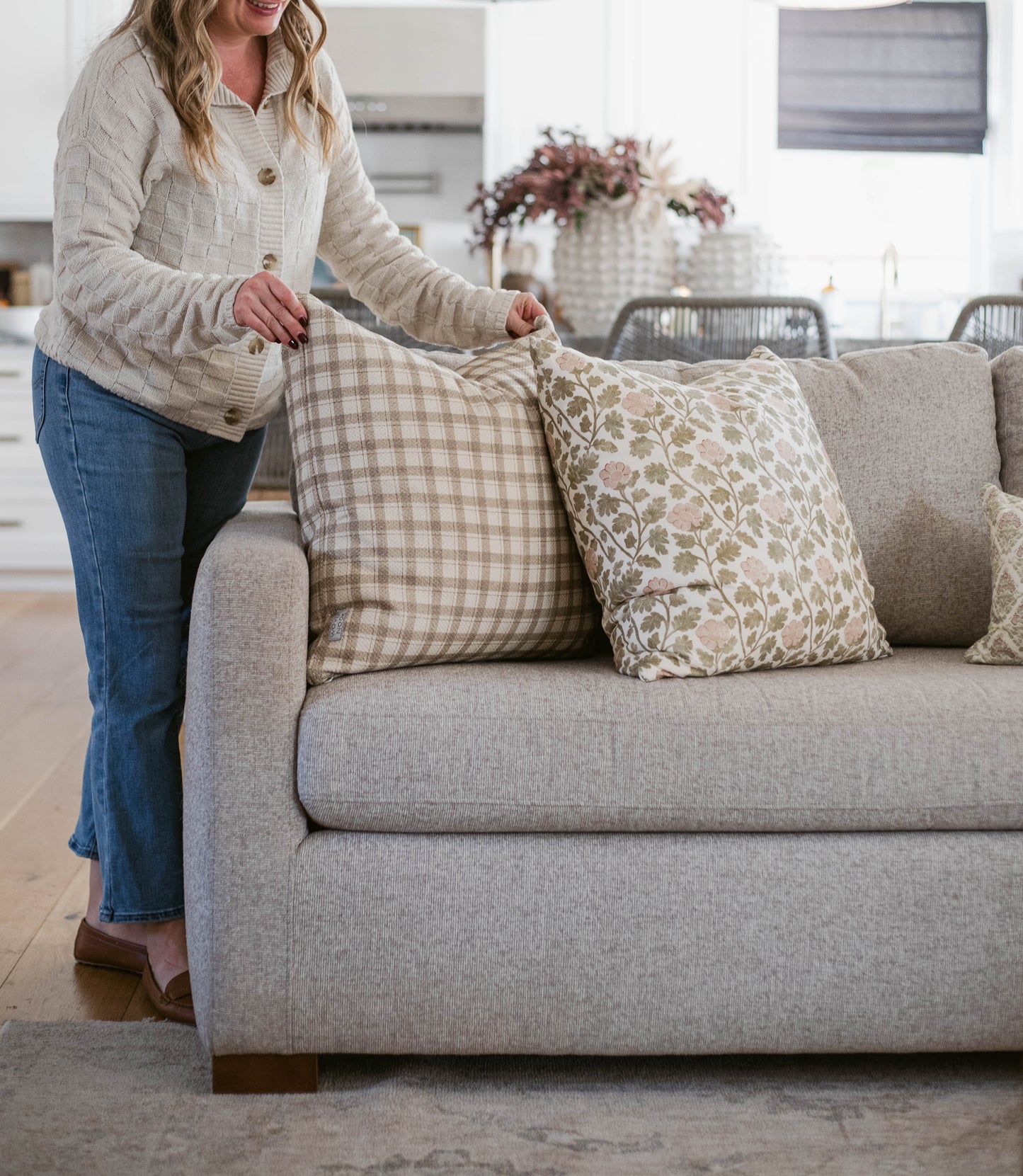 Woman arranging patterned throw pillows on a light gray upholstered sofa in a bright living room.