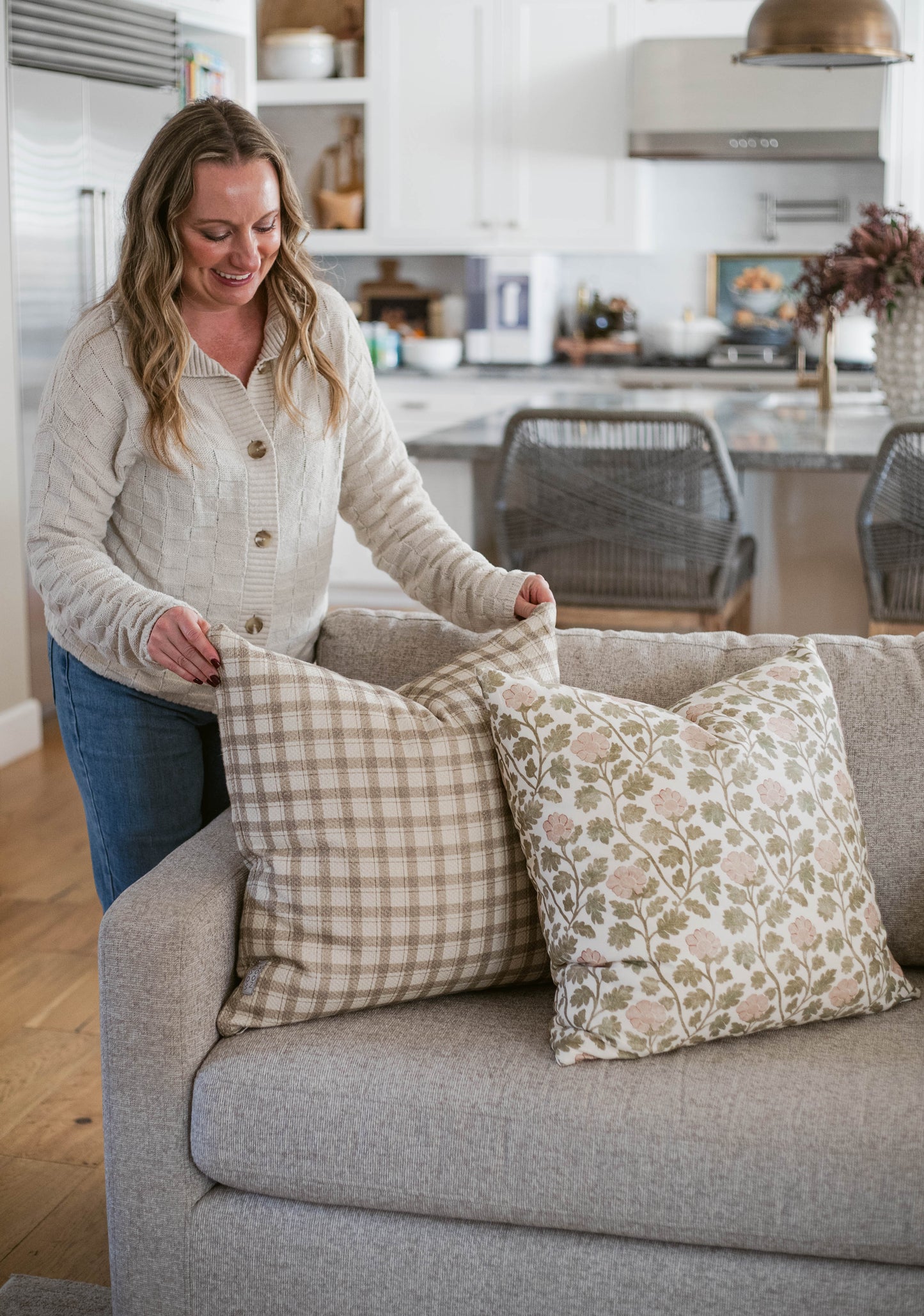 Woman holding two throw pillows on a gray sofa; one with tan gingham pattern, one with pink floral print.