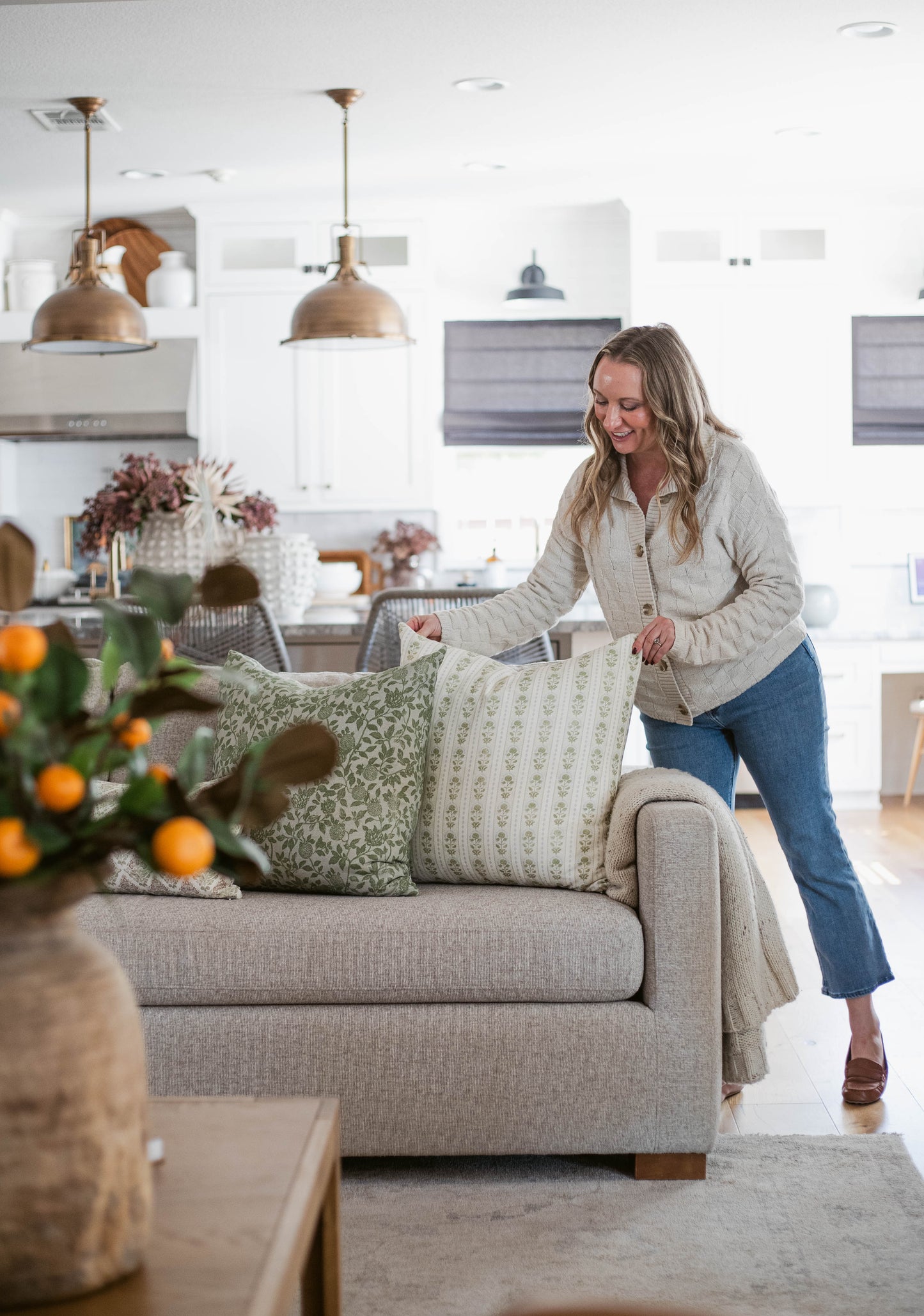 Woman arranging green and cream patterned pillows on a beige upholstered sofa in a bright kitchen-living room.