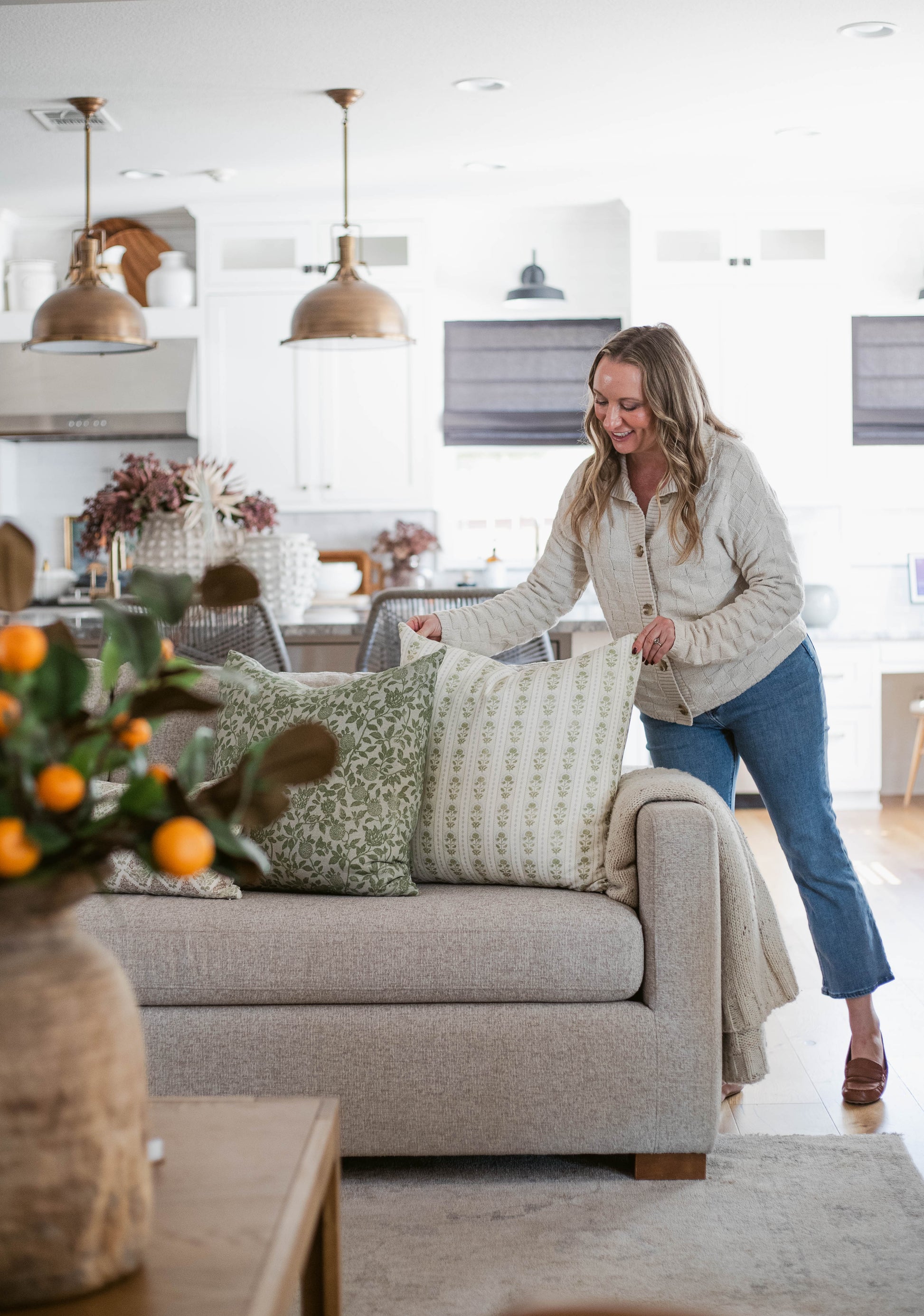 Woman arranging green and cream patterned pillows on a beige upholstered sofa in a bright kitchen-living room.