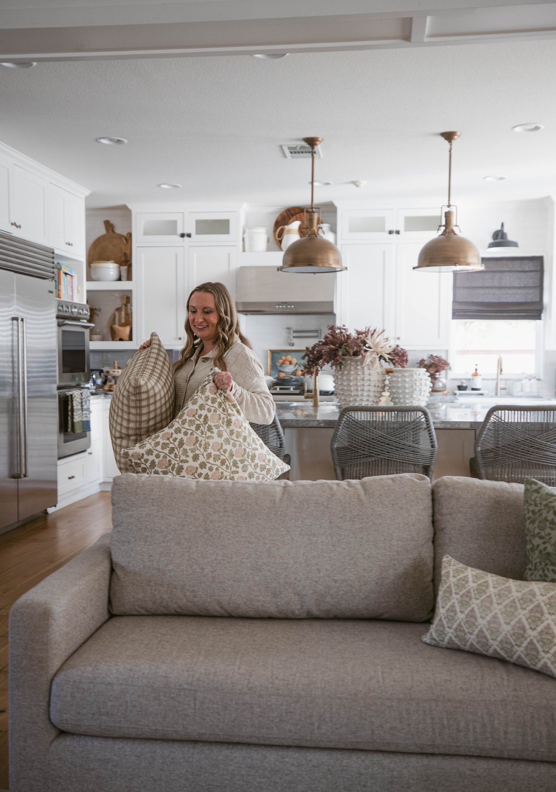 Woman holding patterned and checkered decorative pillows while sitting on a gray sofa in a modern kitchen.