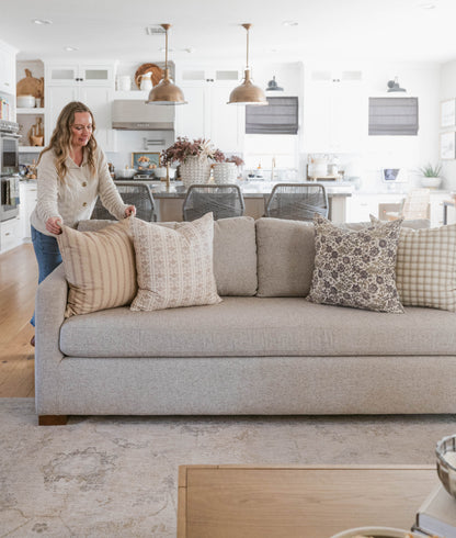 Beige upholstered sofa with five decorative pillows in neutral patterns, displayed in a bright kitchen and living room settin