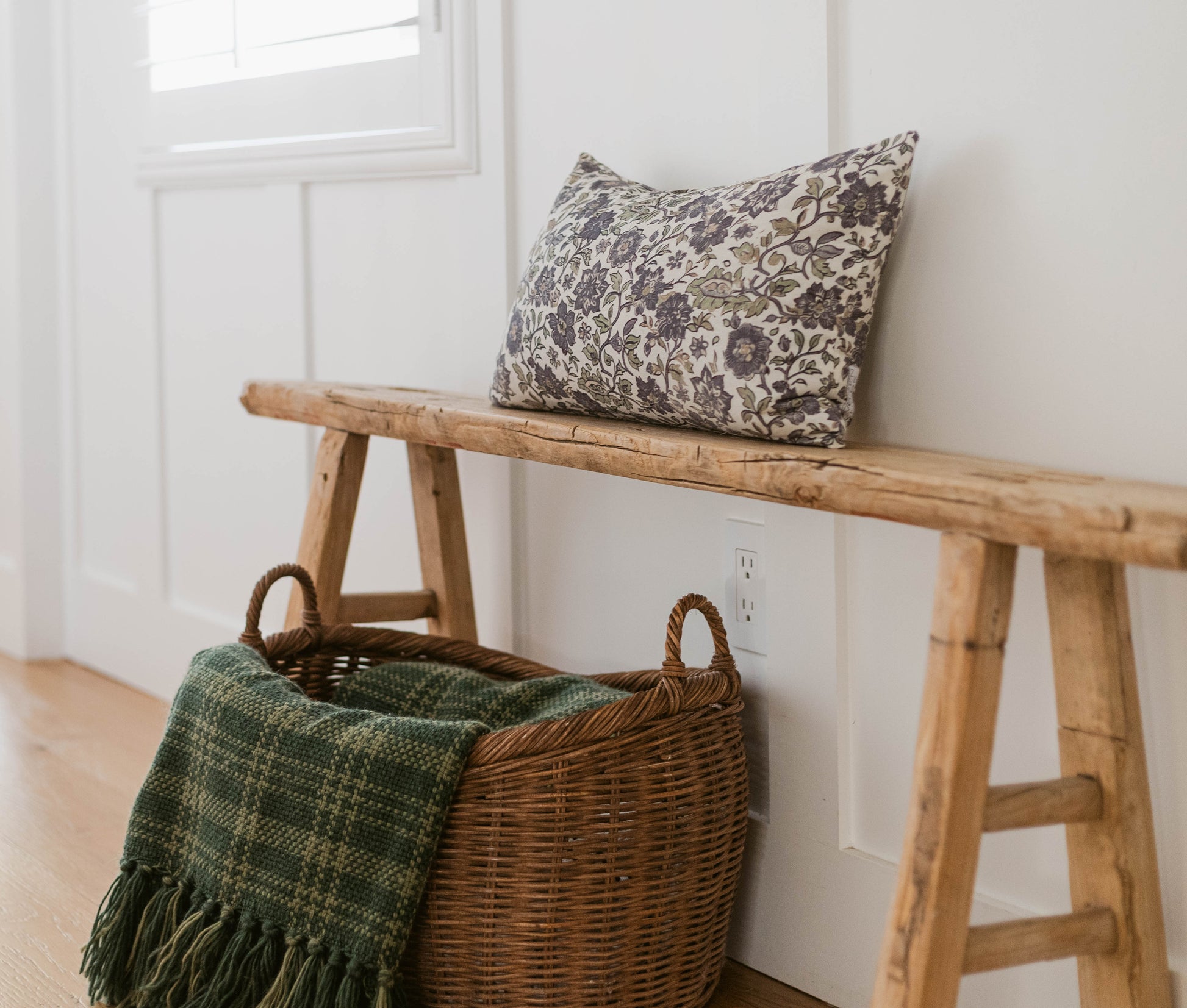 Hattie pillow with gray floral damask pattern displayed on wooden bench next to wicker basket with green throw blanket.