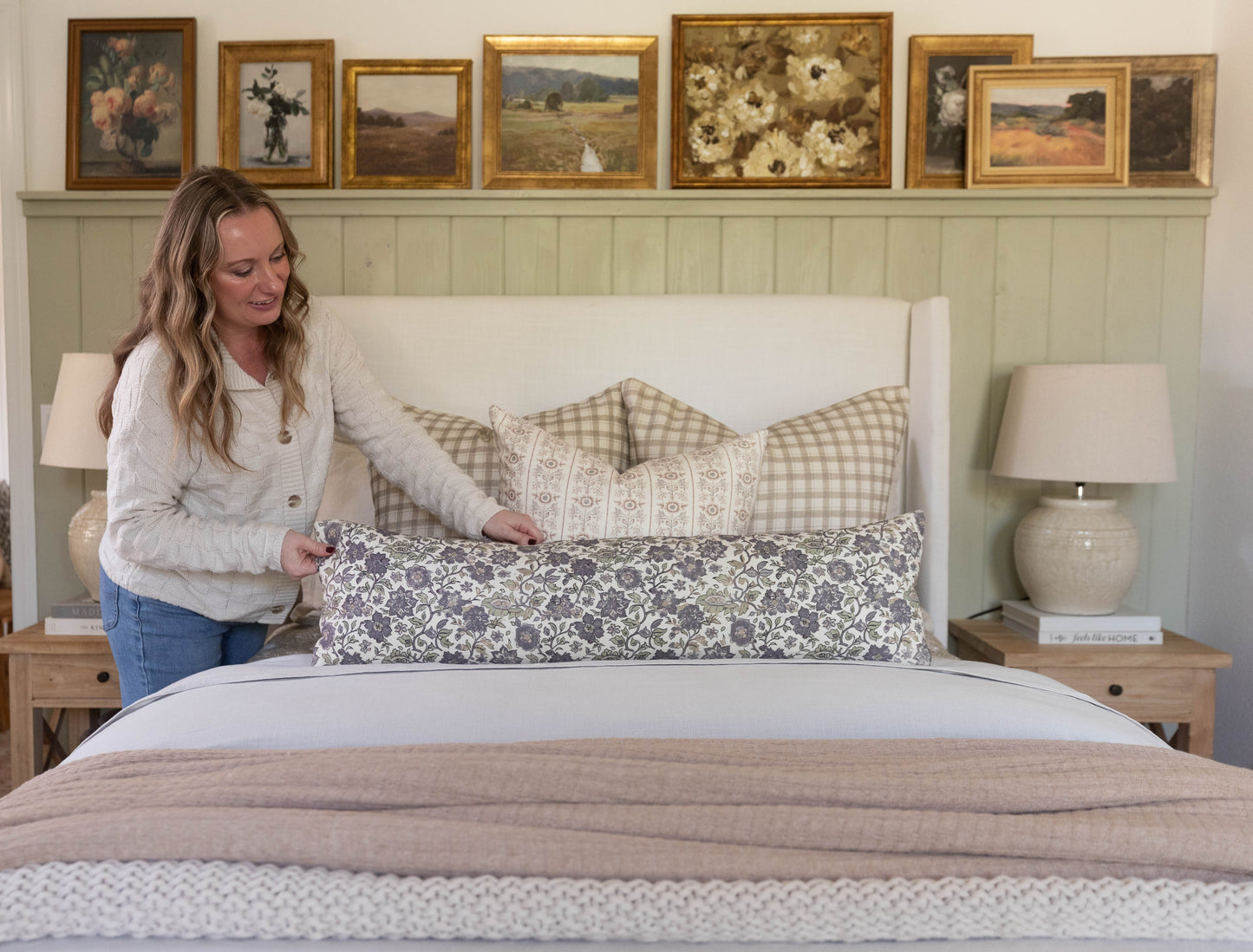 Styled bedroom with cream upholstered headboard, layered pillows in neutral patterns, beige bedding, and framed landscape art
