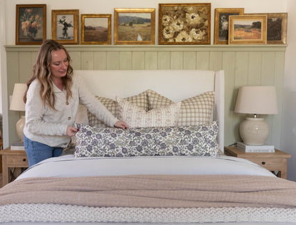 Styled bedroom with cream upholstered headboard, layered pillows in neutral patterns, beige bedding, and framed landscape art