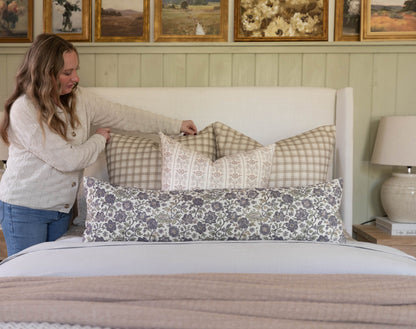 Woman arranging patterned and checkered decorative pillows on a cream upholstered bed headboard.