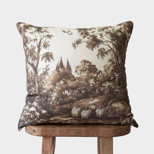 Cream and brown toile pillow with forest landscape and castle illustration, displayed on wooden stool.