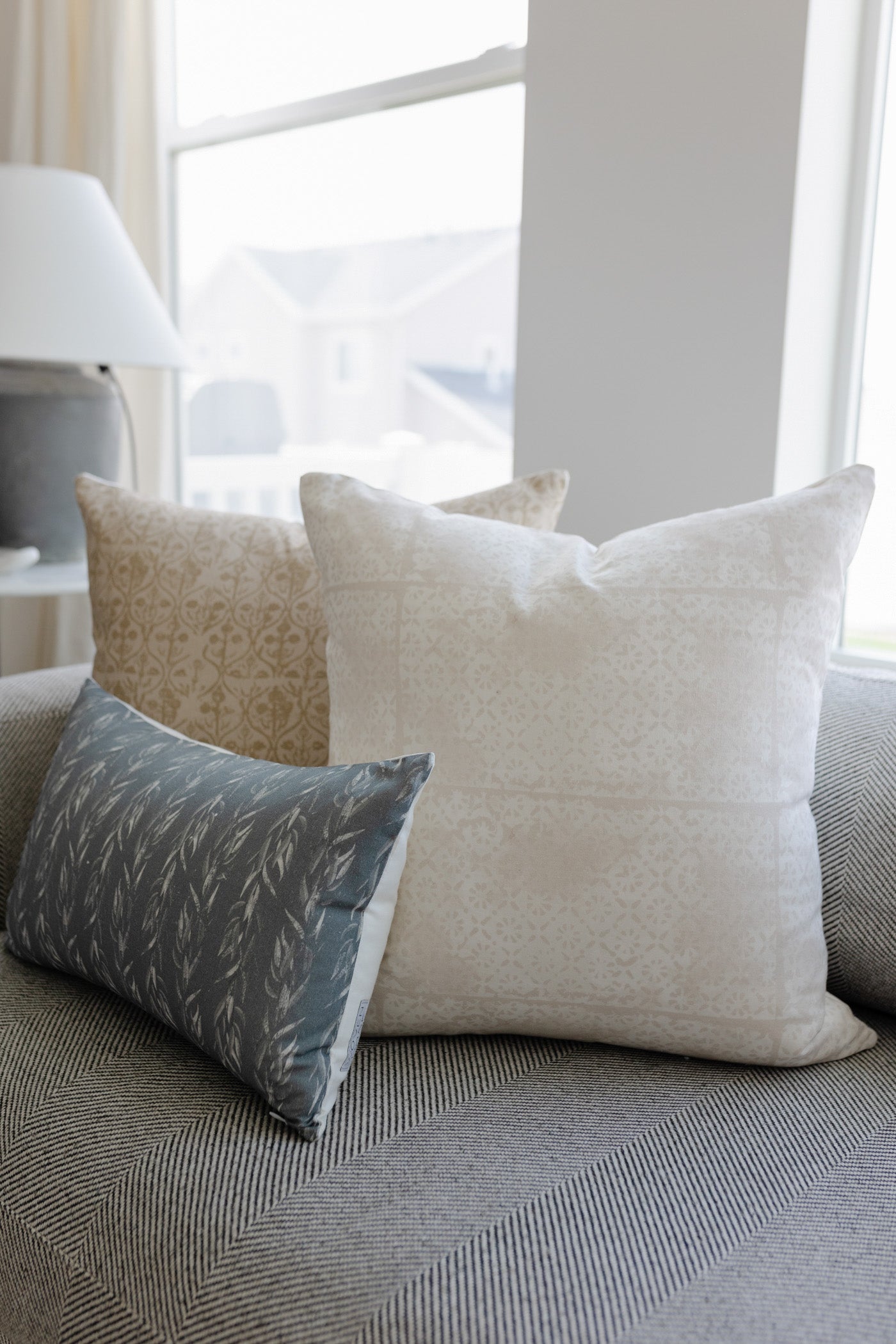 Cream and gray faded floral patterned throw pillow displayed on a gray upholstered sofa by a window.