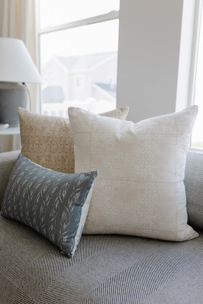 Cream and gray faded floral patterned throw pillow displayed on a gray upholstered sofa by a window.