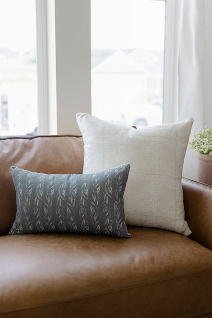 Rectangular pillow with faded floral leaf pattern in gray-blue on tan leather sofa next to cream pillow.