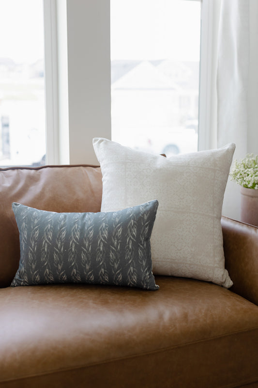 Rectangular pillow with faded floral leaf pattern in gray-blue on tan leather sofa next to cream pillow.