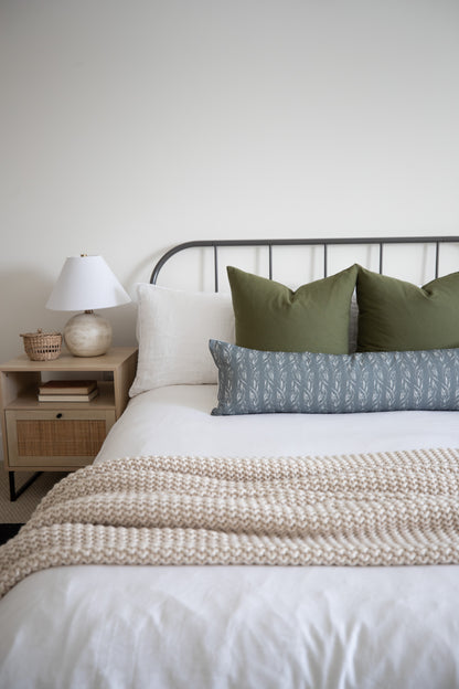 Sage green lumbar pillow with leaf pattern displayed on bed with white and cream pillows.