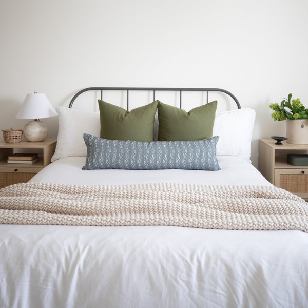 Sage green lumbar pillow with solid fabric displayed on bed between white and patterned pillows.