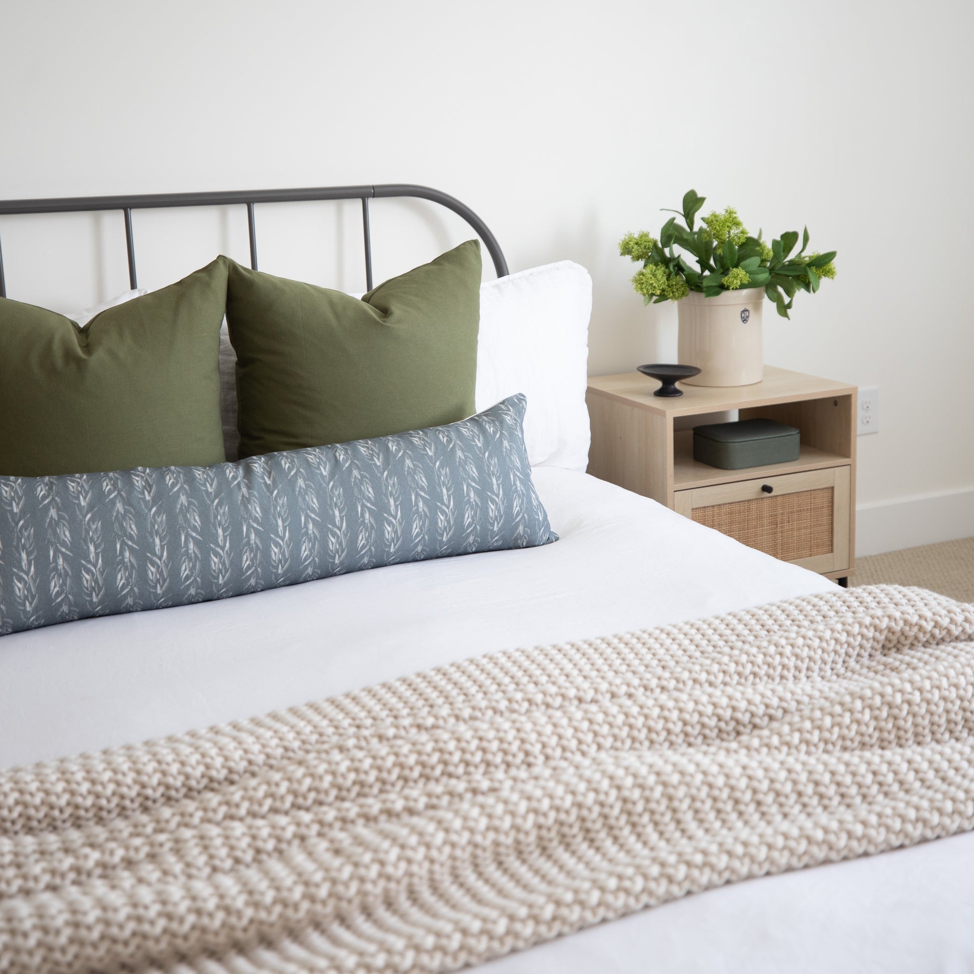 Sage green and blue leaf-patterned lumbar pillow on a bed with white bedding and a metal headboard.