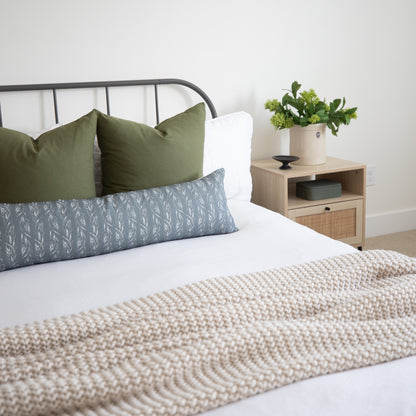 Sage green and blue leaf-patterned lumbar pillow on a bed with white bedding and a metal headboard.