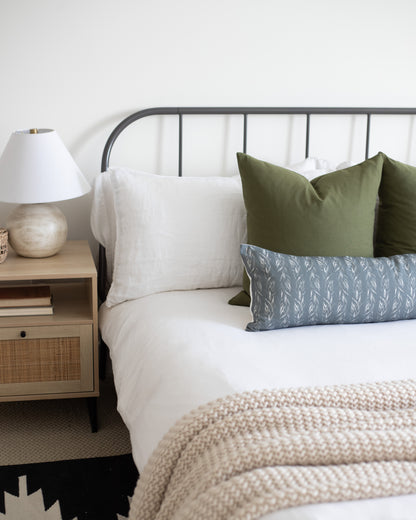 Rectangular lumbar pillow with sage green solid fabric, displayed on bed with white and patterned pillows.