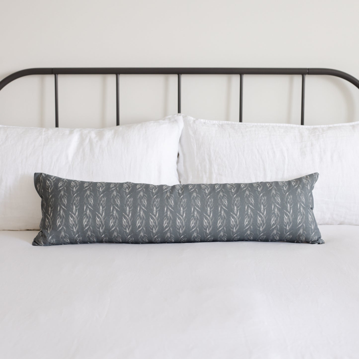 Sage green lumbar pillow with repeating leaf pattern displayed on bed with white pillows and black metal headboard.