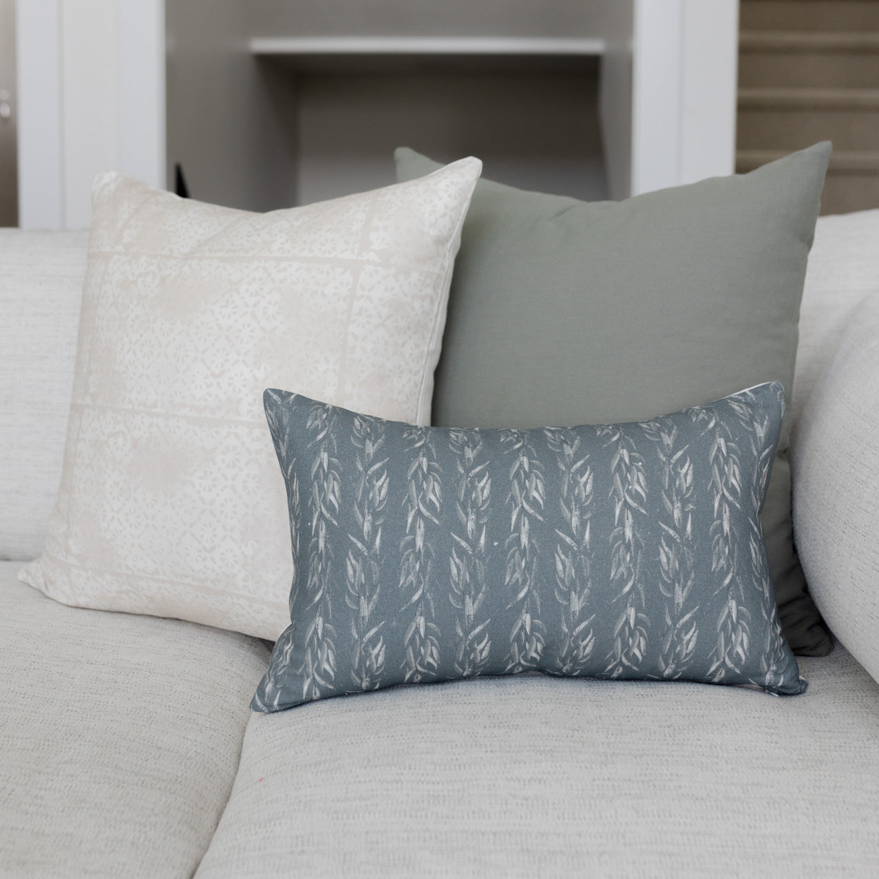 Rectangular pillow with faded leaf pattern in slate blue and cream, displayed on a light gray sofa with coordinating solid pi