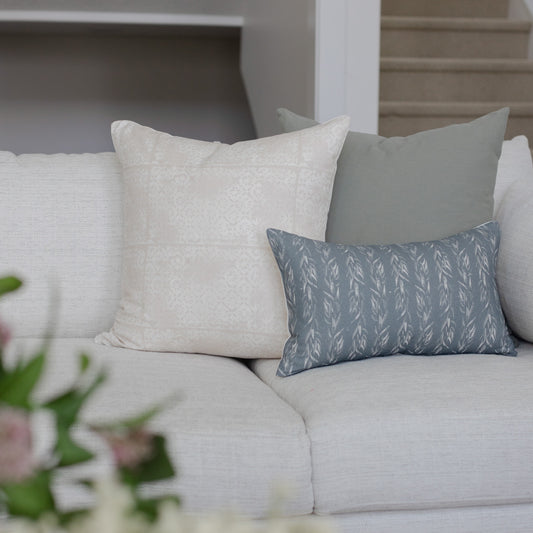 Blue-gray rectangular pillow with faded leaf pattern displayed on light gray sofa with solid pillows.
