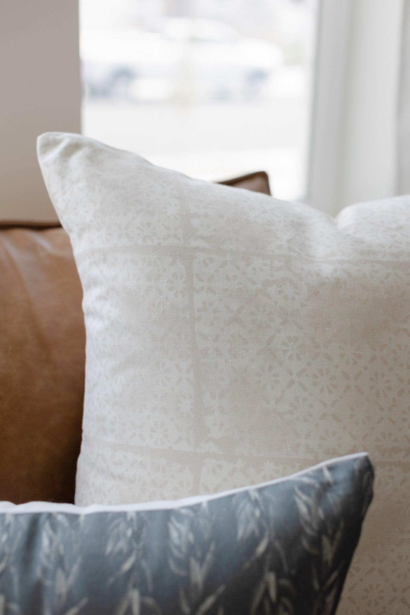 Cream linen pillow with faded floral pattern, displayed on brown leather furniture.