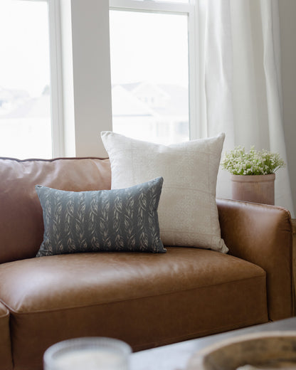 Blue-gray rectangular pillow with faded floral leaf pattern displayed on brown leather sofa.