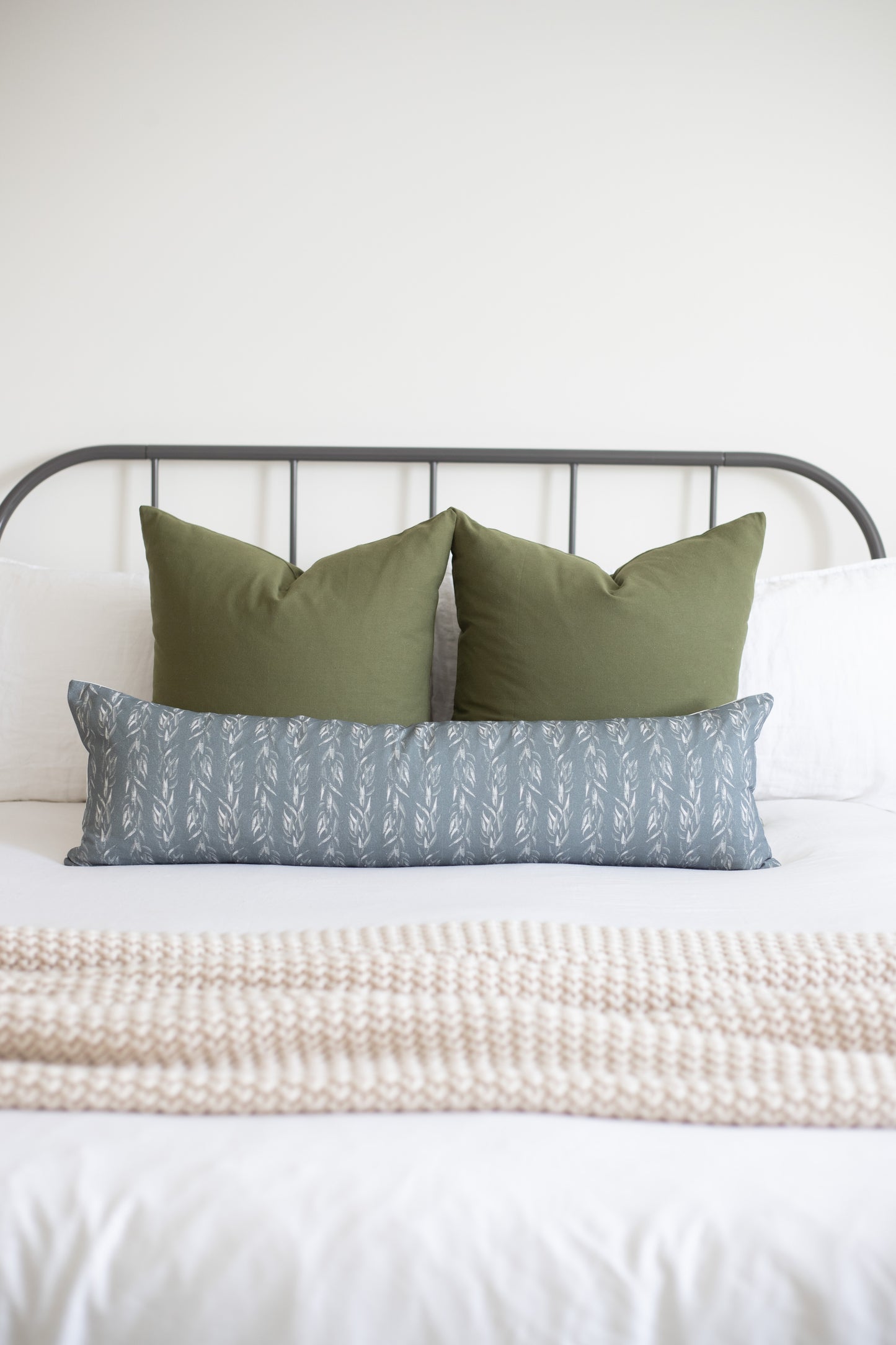 Sage green solid lumbar pillow styled with coordinating leaf-patterned pillow on a metal bed frame.