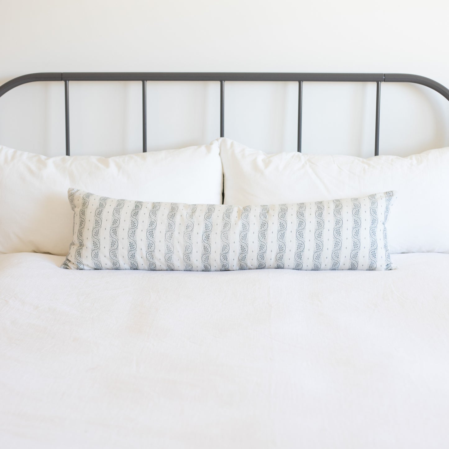 Long rectangular lumbar pillow with gray and white vertical striped pattern on bed with white pillows and metal headboard.